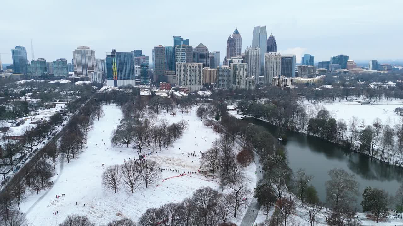 Aerial push in on people sledding in a snow covered Piedmont Park with Midtown Atlanta in the background.