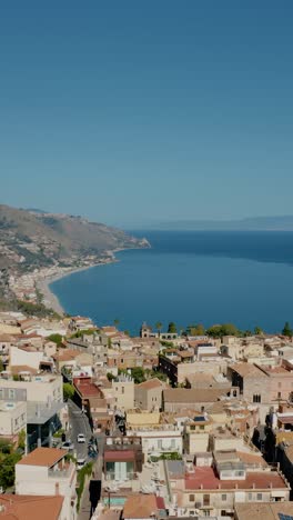 vertical aerial drone shot over the rooftops of ancient town of taormina in sicily. distant view to island's coast line.