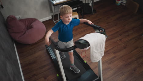 little kid walking on treadmill inside modern room with wooden floor, gripping both handles while looking down, towel placed on treadmill panel, soft beanbag and keyboard setup visible in background