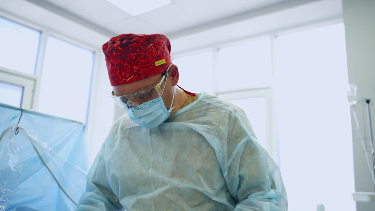 Male surgeon in red cap, mask, glasses and coat. Busy doctor working at operation in modern surgical room.