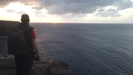 hombre maduro con una mochila en la espalda admira la puesta de sol desde los acantilados de la costa del municipio de gáldar