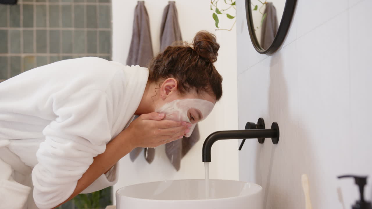 Applying face mask, woman in bathrobe washing face at bathroom sink
