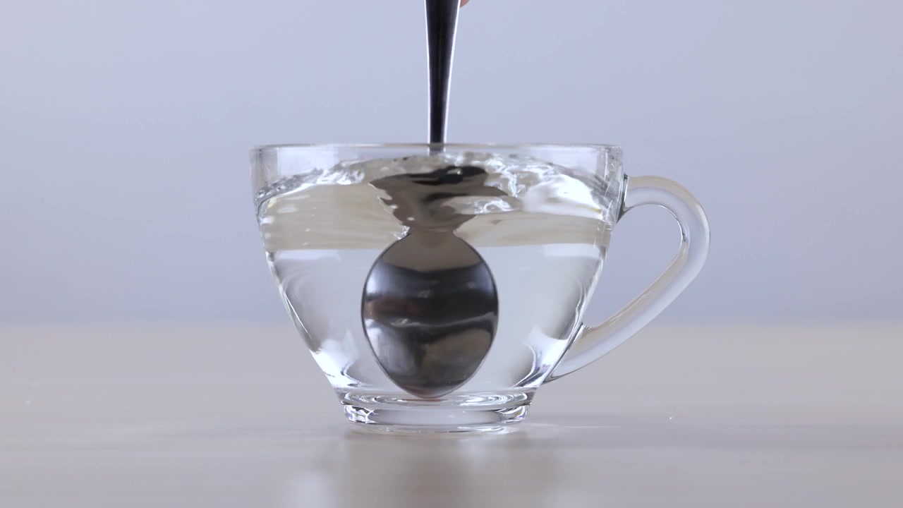 Un cucchiaio di metallo mescola acqua calda in una tazza di vetro trasparente, creando un vortice