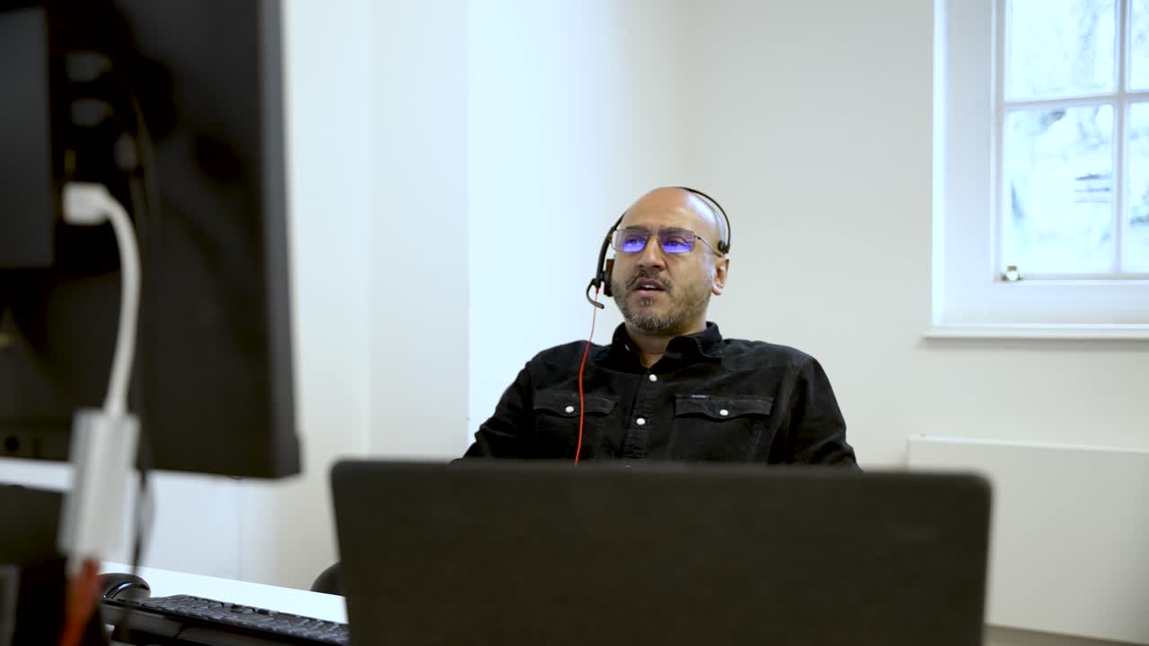 South asian businessman wearing blue light filtering glasses talking on a headset in a modern office, office swiveling on chair