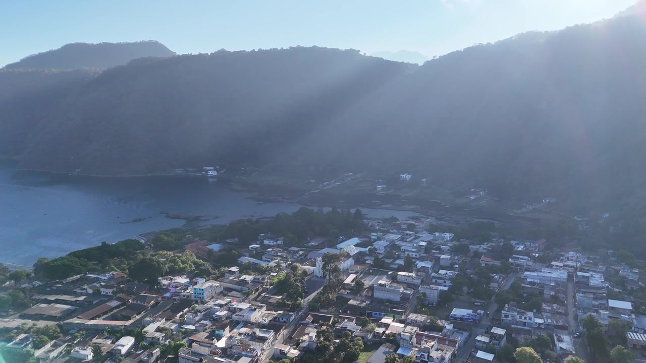 Pemandangan udara drone di atas San Lucas Guatemala saat perairan, permukiman tropis, langit cerah, pohon palem, dan pegunungan berapi berhutan terlihat, menampilkan lanskap yang semarak