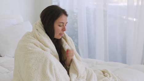 sick woman wrapped in a white blanket coughing sitting on the bed at home