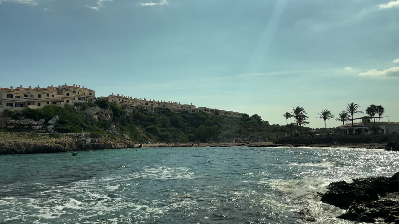 Olas brillando bajo la luz del sol cerca de palmeras y costa rocosa en la isla de Mallorca, España
