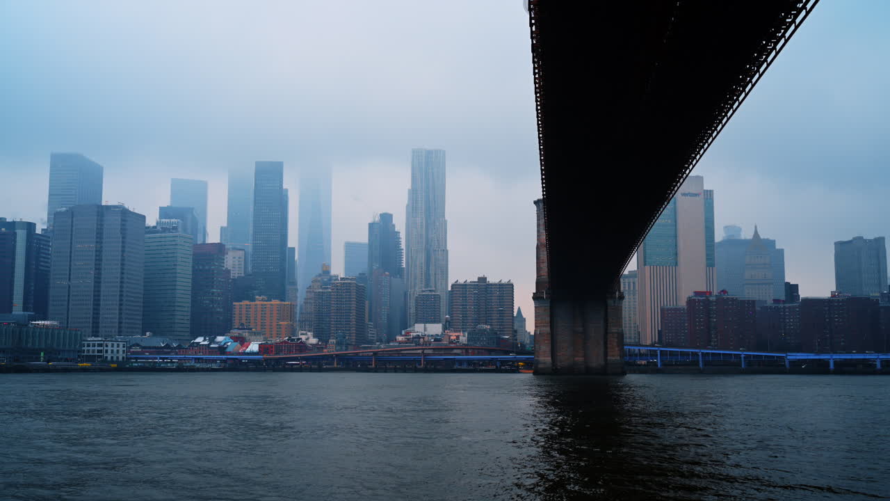 New York, USA, 9 February 2026: Lower Manhattan skyline view from under the bridge. Foggy cityscape featuring skyscrapers and water seen from beneath a bridge structure