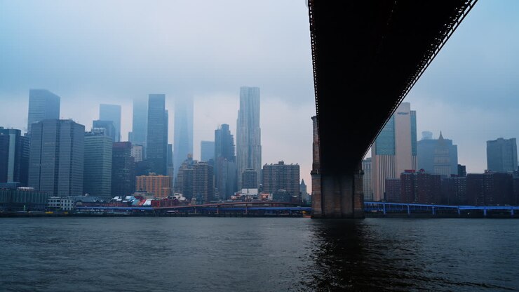 New York, USA, 9 February 2026: Lower Manhattan skyline view from under the bridge. Foggy cityscape featuring skyscrapers and water seen from beneath a bridge structure