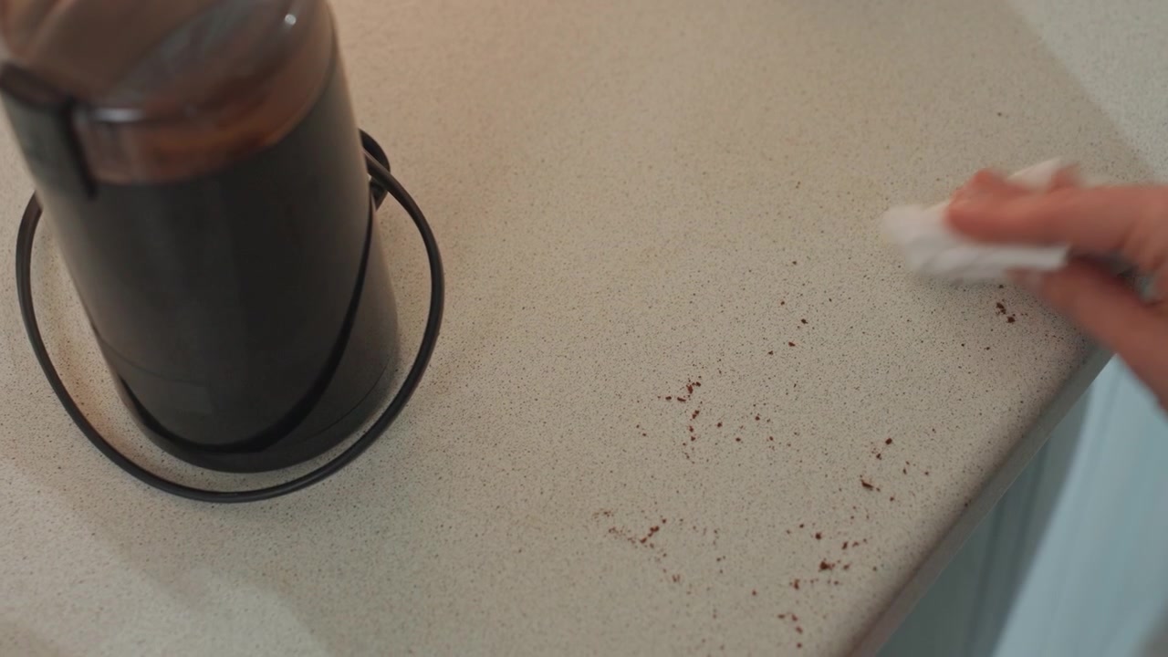 Primo piano di fondi di caffè sul bancone, mano che pulisce con un panno rimuovendo i fondi sparsi, macinacaffè elettrico nelle vicinanze, rapida pulizia, luce mattutina brillante, pulizia pratica, superficie testurizzata, gesto efficiente.