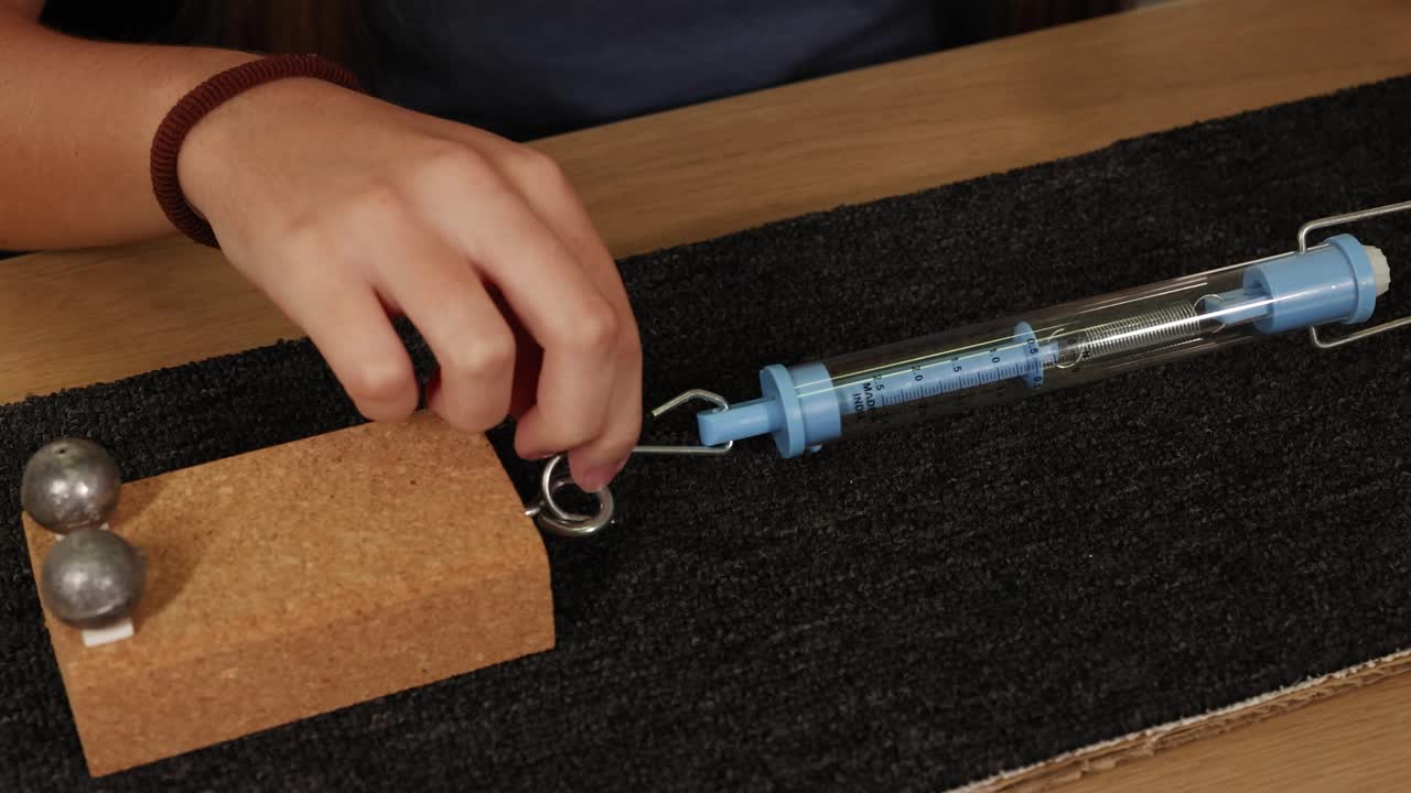 Hands using a spring scale to measure force on a block, illustrating friction on a carpeted surface