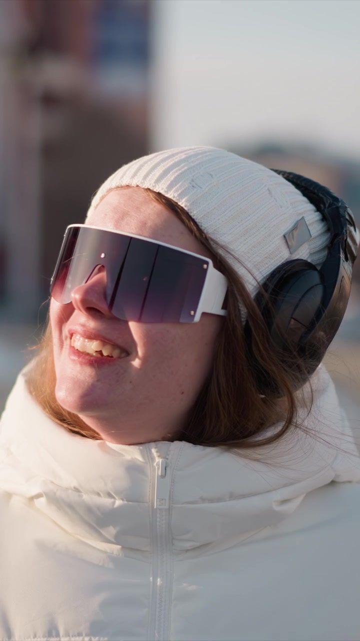 Woman wearing white knit hat large dark sunglasses and black headphones smiles outdoors in bright sunlight. Woman is dressed in thick padded winter jacket with visible texture details