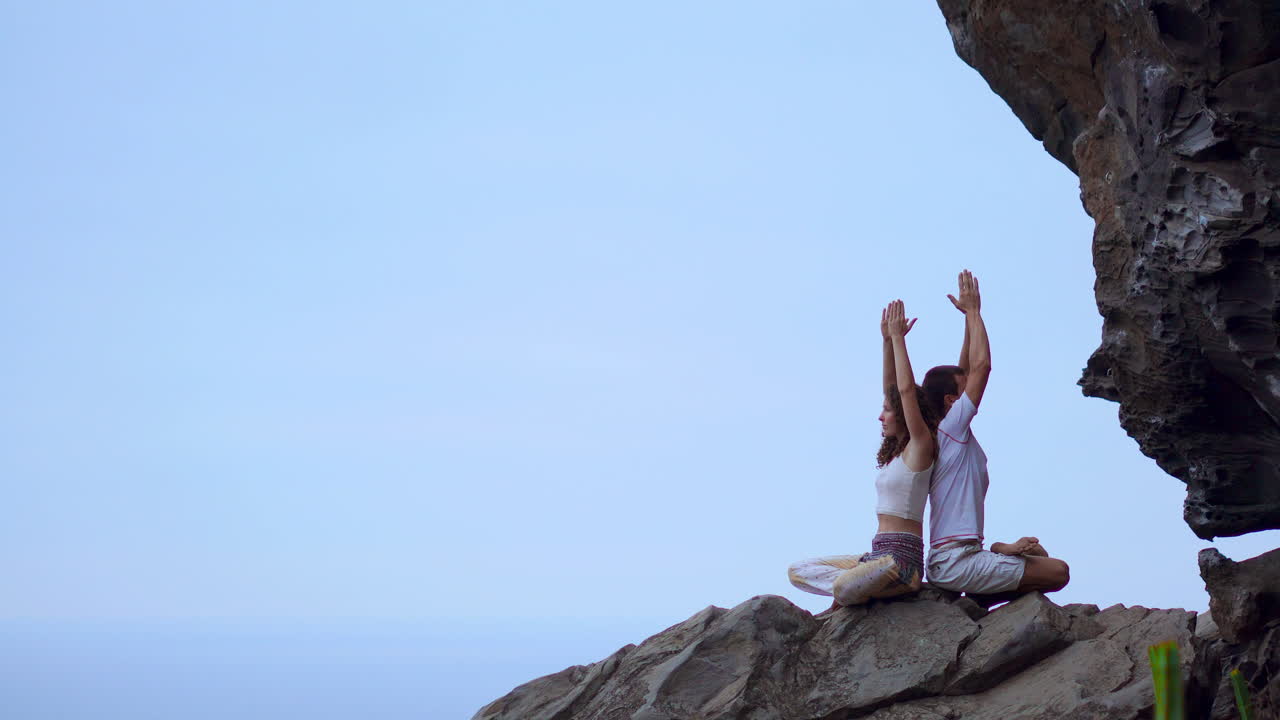 On a mountain's peak, a man and woman sit back to back on a rock, engaging in meditation and yoga with the ocean as their backdrop