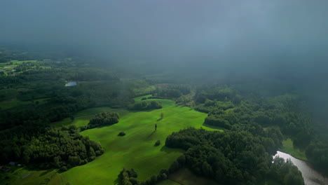 A-High-Angle-Shot-Of-A-Green-Grassy-Landscape-With-Forests-And-Ponds-Below-Fluffy-Clouds