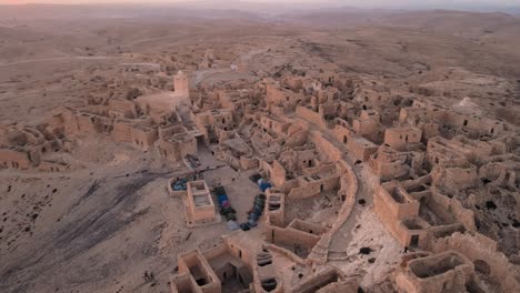 a desert city with ancient ruins and structures, including buildings and rocky outcrops. the sun is setting over the landscape, casting warm light on the area. location: tunisia, gabes , zraoua.