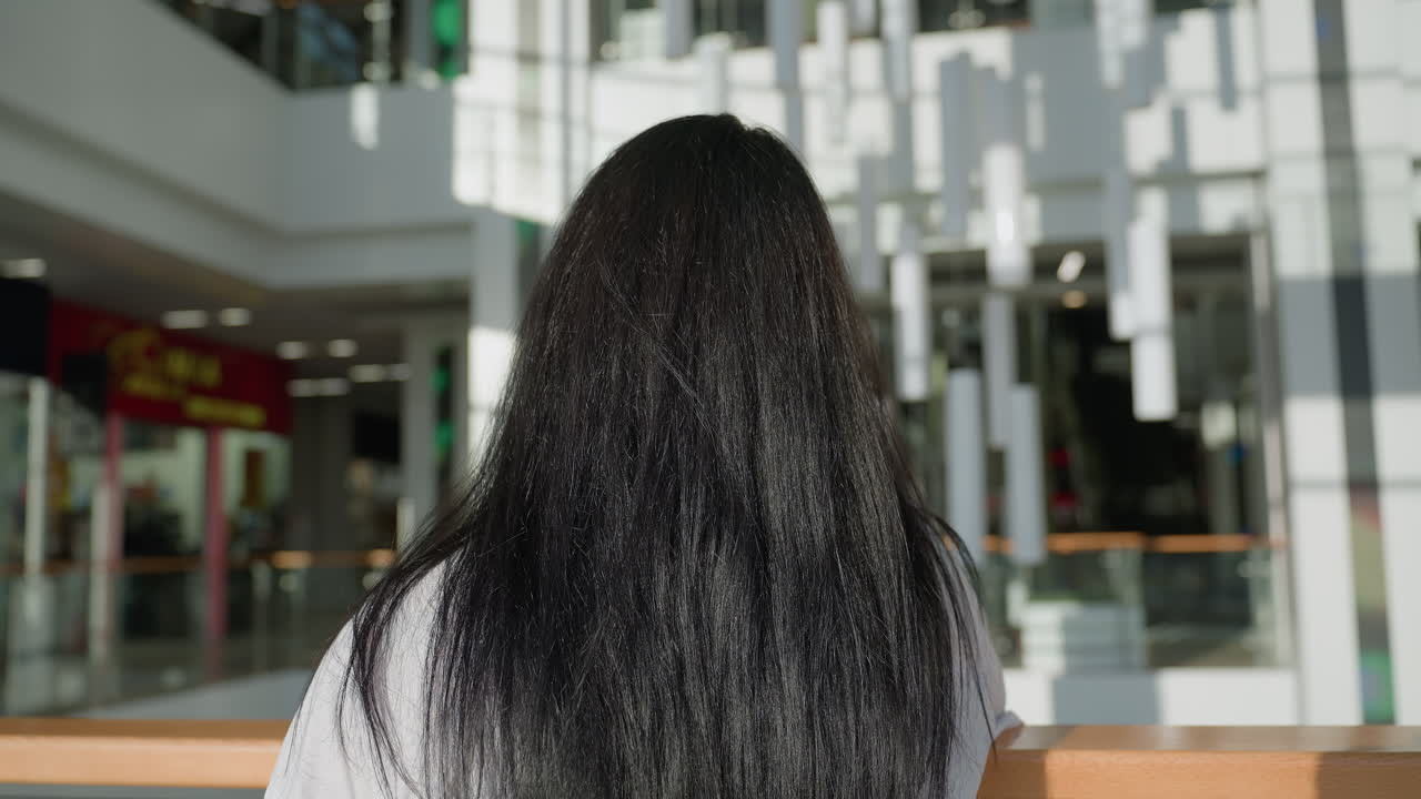 Back view of woman with long dark hair resting on rail inside bright modern mall with contemporary interior design, decorative hanging lights, and glowing reflections in spacious atmosphere