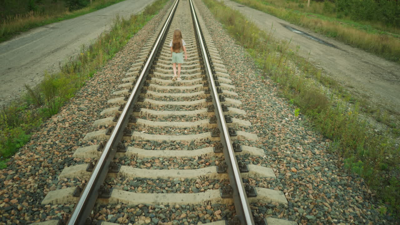 Rear view of girl walking confidently on railway track in rural setting holding object in hand, surrounded by gravel and overgrown greenery, with long hair and casual outfit under natural daylight