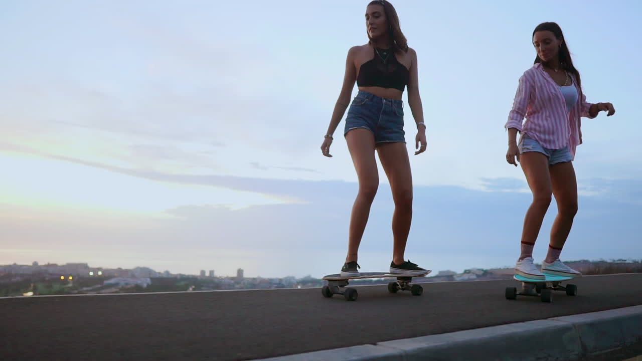 In slow motion, two friends wearing shorts ride skateboards along a road at sunset, with mountains and a captivating sky as the backdrop