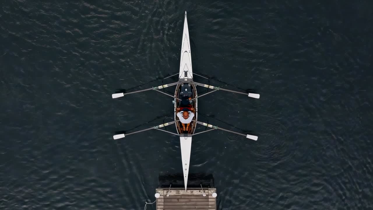 Pushing off dock, two rowers sculling double scull into open water, making ripples, in orange PFD