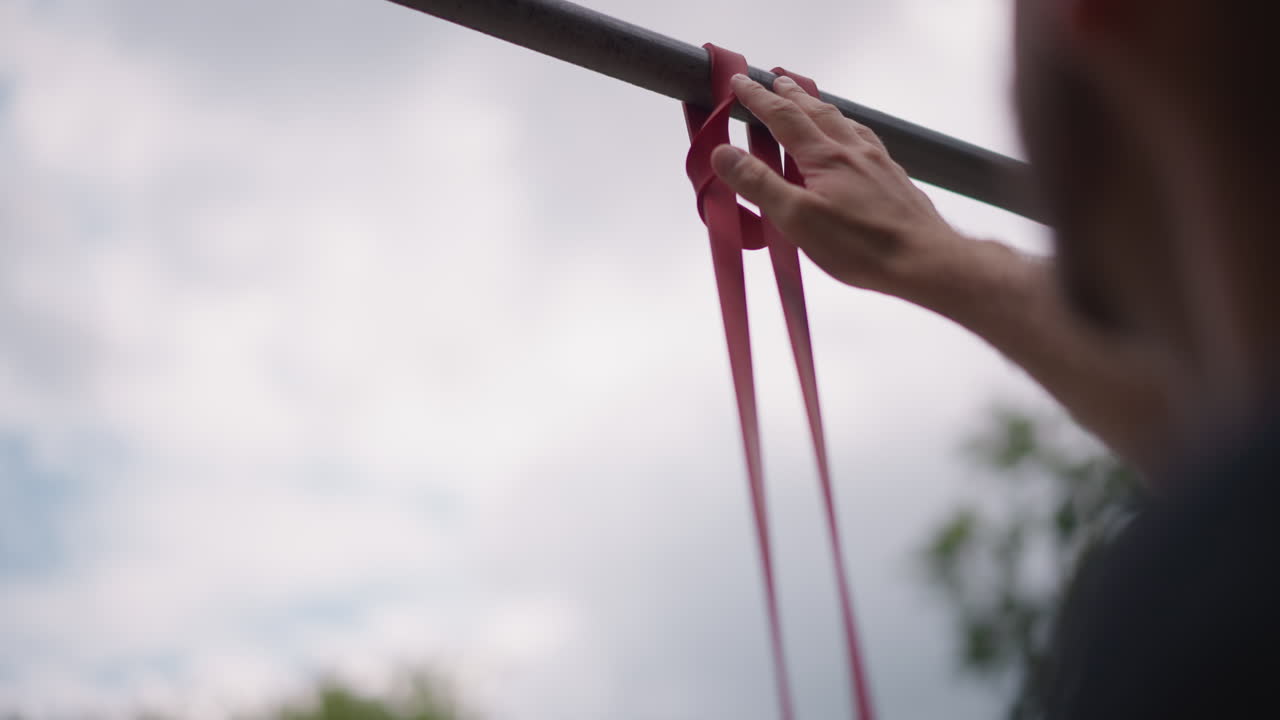 Person Carefully Fastening Loop Onto Overhead Bar, Man Meticulously Securing Red Band Around Pullup Bar Overhead, Individual Diligently Tying Loop Around Metallic Bar Under Cloudy Sky For Safety
