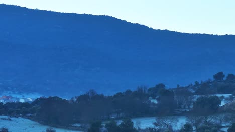 Vuelo-Lateral-Con-Un-Dron-En-Un-Amanecer-Helado,-Hay-Ese-Color-Azul-Turquesa-En-El-Rodaje,-Visualizando-Primero-Un-Prado-Con-árboles,-Luego-Un-Pueblo-Y-Al-Fondo-Una-Montaña-En-Cámara-Lenta