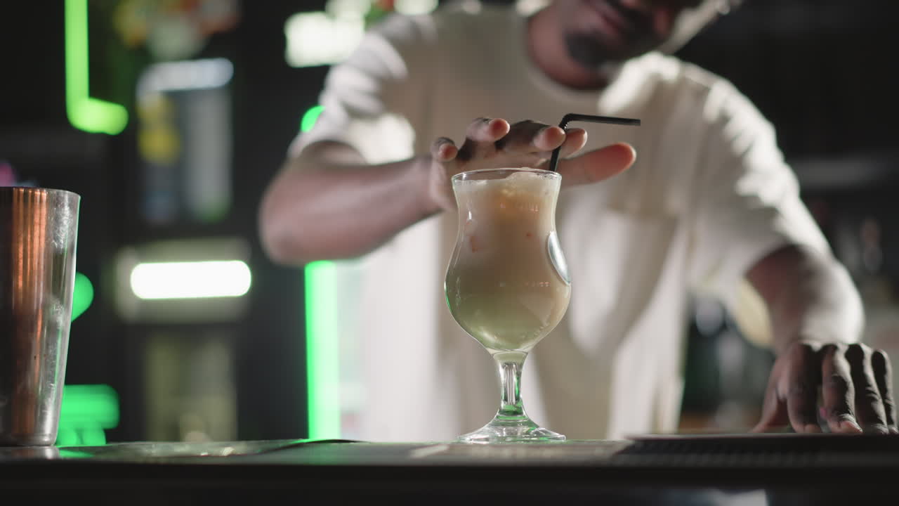 Bartender stirring cocktail in glass with straw, preparing the drink in a vibrant bar. Behind, illuminated Heineken neon sign and bar shelves filled with liquor bottles
