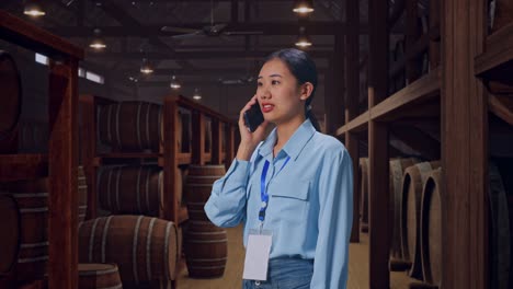side view asian female professional worker standing in cellar of winery, she pick the phone up and started to specking to it with a slicely smile