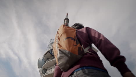 Frog's-eye-view-of-a-radio-tower,-young-hiker-with-an-orange-backpack-walking-pass-the-tower