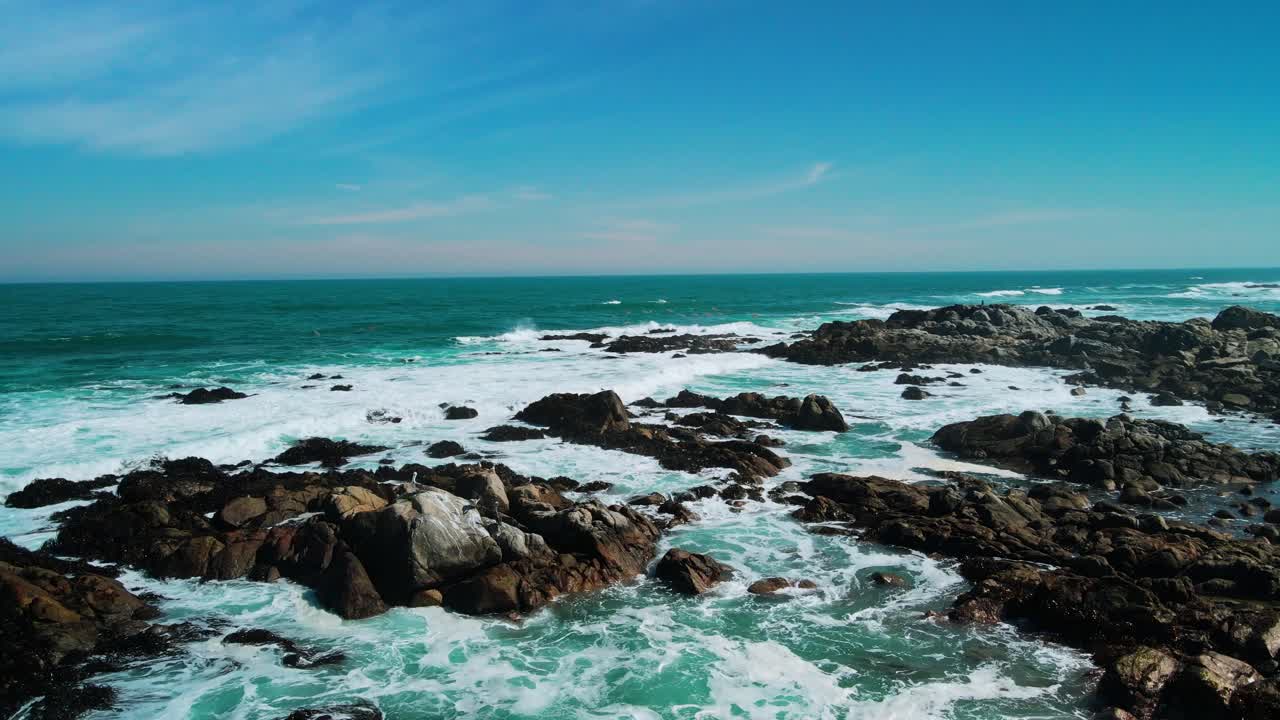 Ripresa orbitale che circonda massi scuri sparsi con onde turchesi spumeggianti che si infrangono tra le rocce e l'Oceano Pacifico aperto che si estende fino all'orizzonte. El Tabo, Cile.
