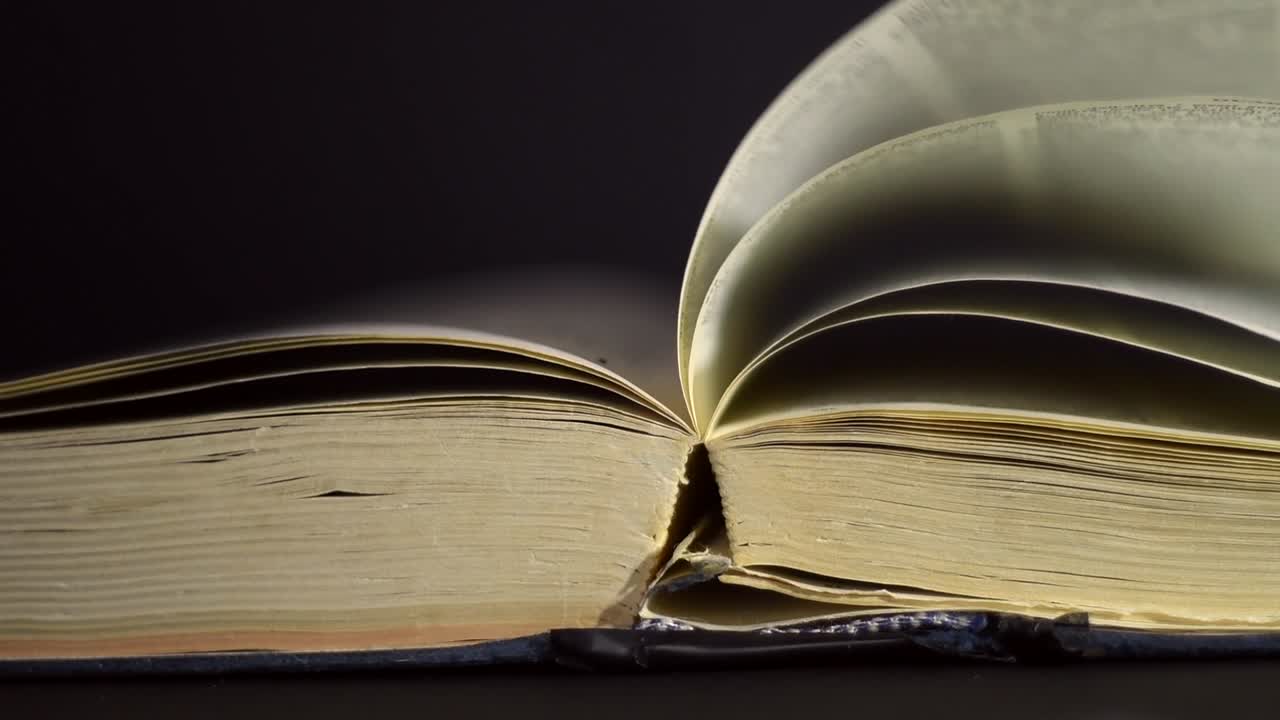 Slow motion, close up of an old, vintage, open book with pages turning on the wind, on a dark room with few light, with black surroundings
