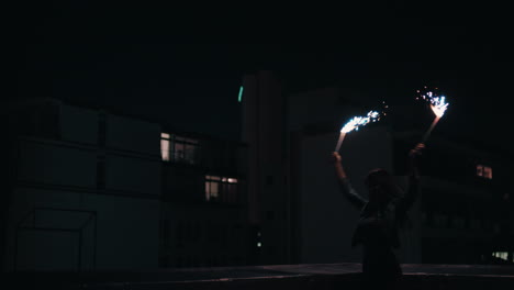 Feliz-Joven-Sosteniendo-Bengalas-Bailando-En-La-Azotea-Por-La-Noche-Celebrando-La-Víspera-De-Año-Nuevo-Disfrutando-De-La-Celebración-Navideña