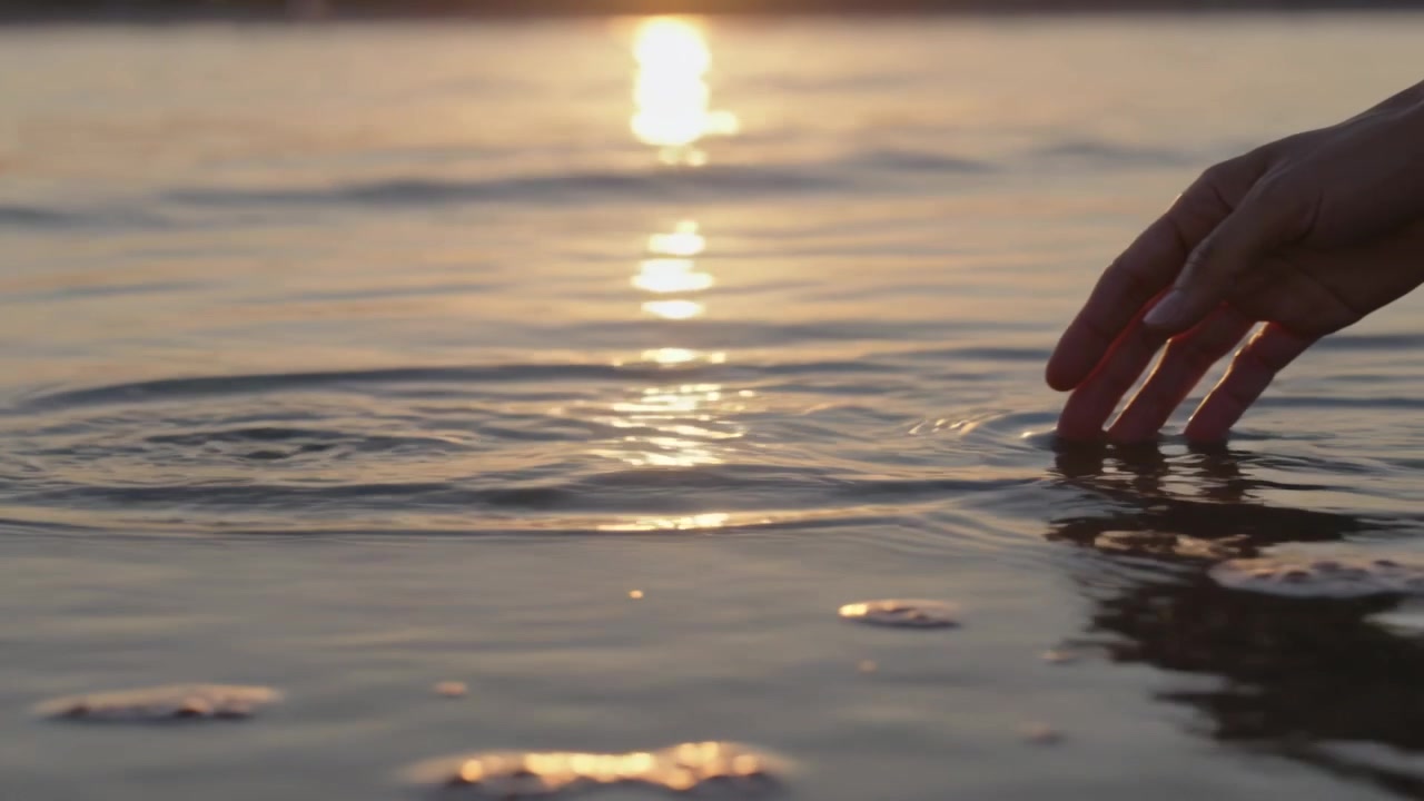 Mano destra che tocca l'acqua, creando increspature sulla riva con la schiuma, distorcendo il bagliore del sole, con spazio per il testo.