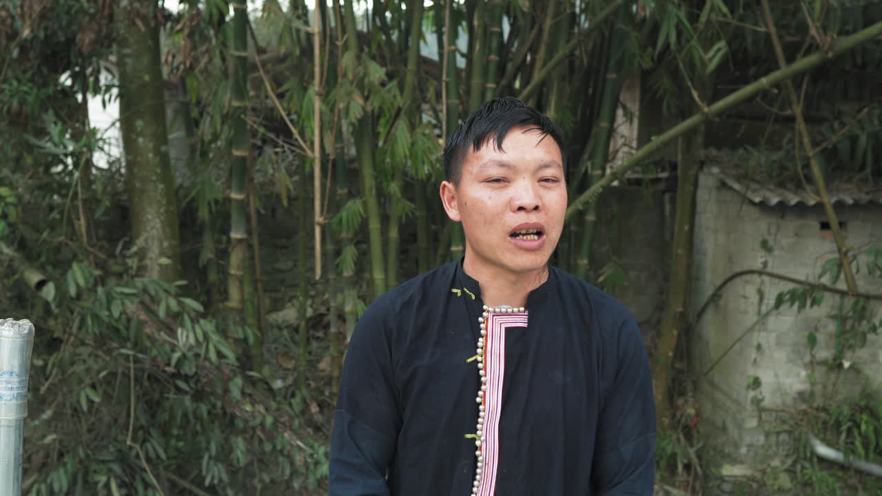 A young Black Dao man speaks in front of bamboo and a small shelter in Tả Phìn, Sa Pa.