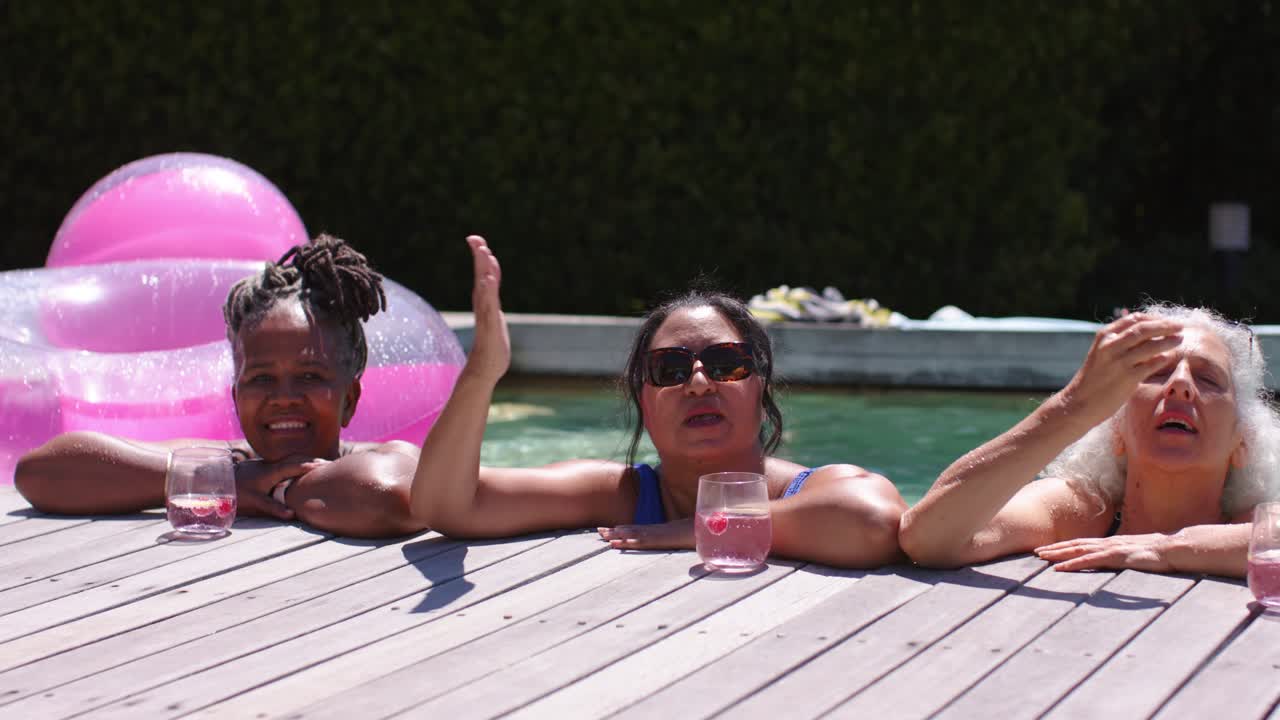 Diverse female friends talking and leaning on wooden deck at backyard pool, sipping pink drinks