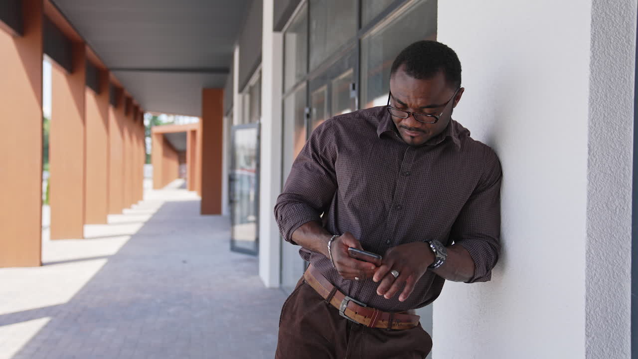 Black guy with sportive body uses mobile phone. Handsome african american man in glasses, stylish shirt standing near office building outdoors using phone.