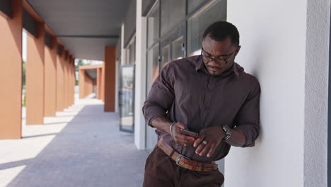 black guy with sportive body uses mobile phone. handsome african american man in glasses, stylish shirt standing near office building outdoors using phone.