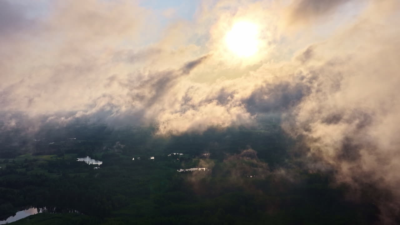 Drone flies through a layer of soft, hazy clouds, moving towards the bright sun, which illuminates the atmospheric scene and the vast, green Latvian landscape visible below at sunset