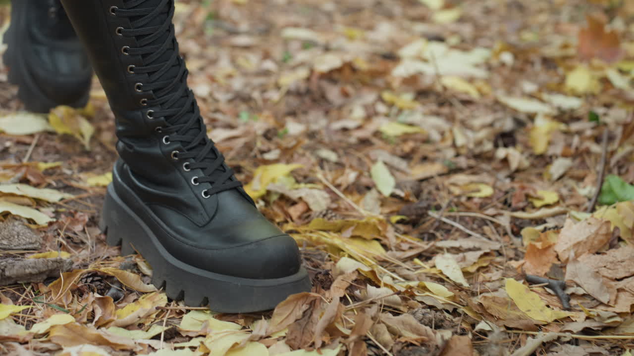 Schwarze Lederstiefel auf Herbstlaub, Frau tritt auf moosigen Baumstumpf, geschnürte Plateausohlen zerquetschen trockenes Laub, stimmungsvolles Waldlicht, körnige Textur, alternative Modeeinstellung, filmreife Nahaufnahme