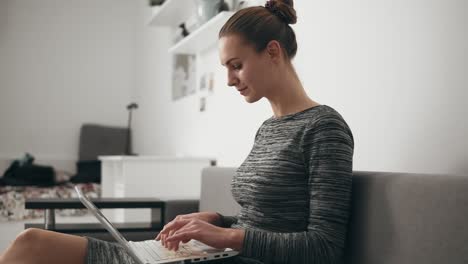 Side-view-of-young-woman-at-home-sitting-on-the-sofa,-working-with-a-laptop-and-typing-text-fast-looking-at-the-screen