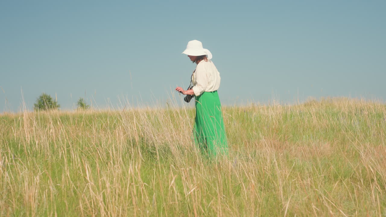 Rear view of woman wearing wide white hat and green skirt walking through dry grassy field under open blue sky, holding camera and observing natural surroundings