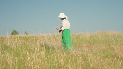 rear view of woman wearing wide white hat and green skirt walking through dry grassy field under open blue sky, holding camera and observing natural surroundings