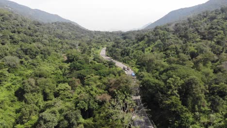 Aerial-flyover-indian-asphalt-highway-with-traffic-surrounded-by-green-nature-forest-mountains-during-sunny-day