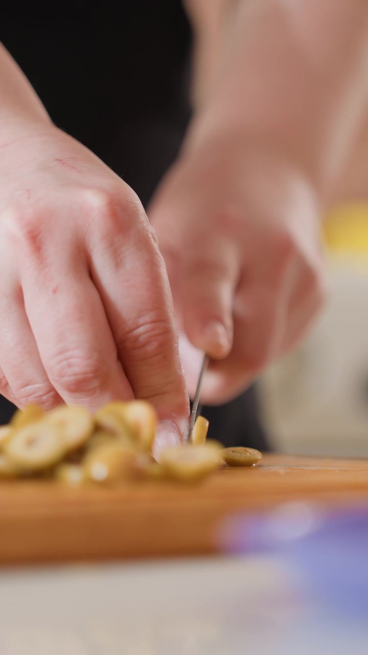 Primo piano frontale basso che mostra mani che affettano piccoli pezzi di cibo rotondi in una pila ordinata su un tagliere di legno, con un colpo di coltello controllato verso il basso, presa ferma, polpastrelli arricciati e una calda luce interna.