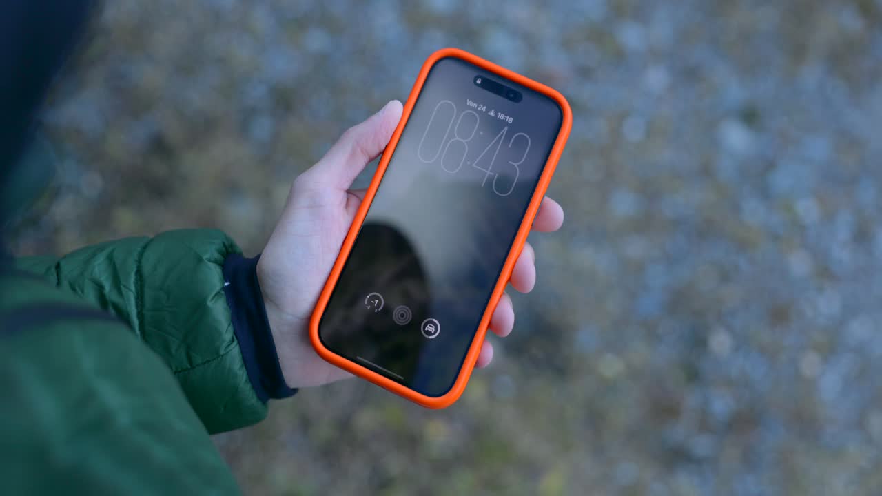 A person in a green jacket holds an orange rugged smartphone outdoors, inserting a SIM card while the device is visible on a nature trail