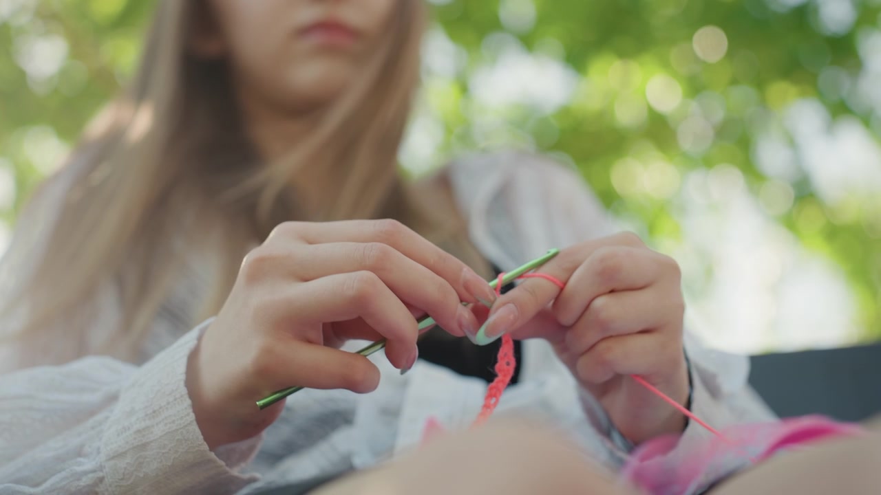 Close up of young woman crocheting with pink yarn and green hook in soft natural light. Her hands manipulate materials showing detailed finger movements against outdoor backdrop with blurred