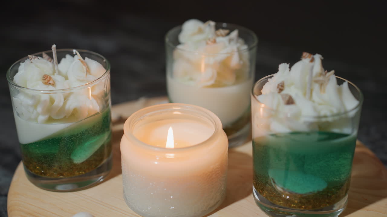 Three transparent glass cupcakes topped with whipped cream decor placed on wooden table beside glowing burning candle, bright soft light focused on desserts creating cozy warm atmosphere