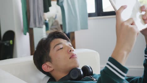 asian boy playing games on digital tablet lying on the couch at home