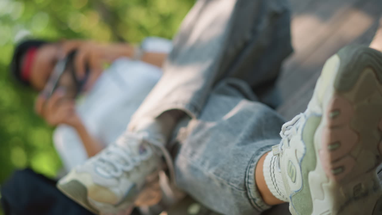 공원 벤치에서 모바일 게임을 하는 아시아 십 대, 발과 운동화가 보이고 나무 판자에 기대어 앉아 햇빛이 비치는 나무와 부드러운 보케, 편안하게 집중하여 휴대폰을 들고 있는 모습은 캐주얼 게이머를 연상시켜요.