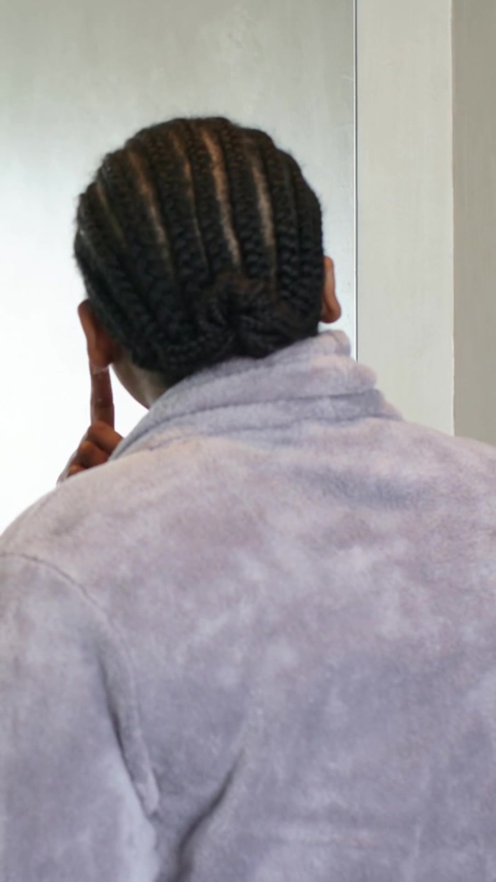 Vidéo verticale : Femme en peignoir avec un chignon tressé se regardant dans le miroir à la maison, touchant son menton et examinant sa peau.