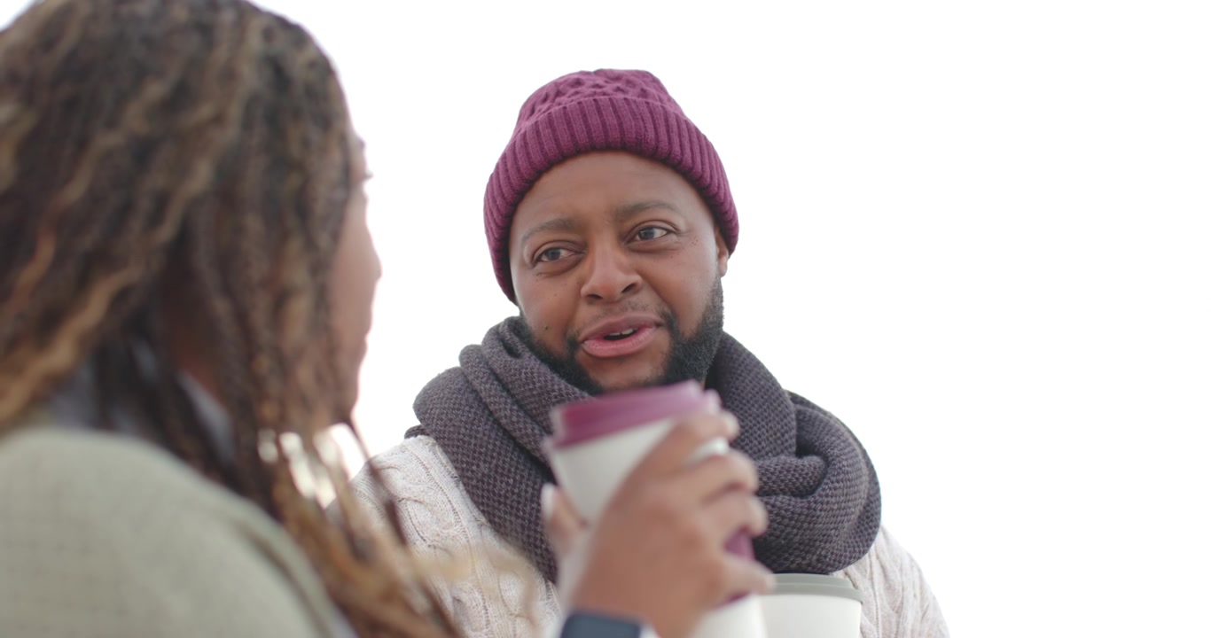 Coppia afroamericana all'aperto sorseggia da bicchieri da asporto, l'uomo con un cappello bordeaux lancia un'occhiata e parla in modo colloquiale.